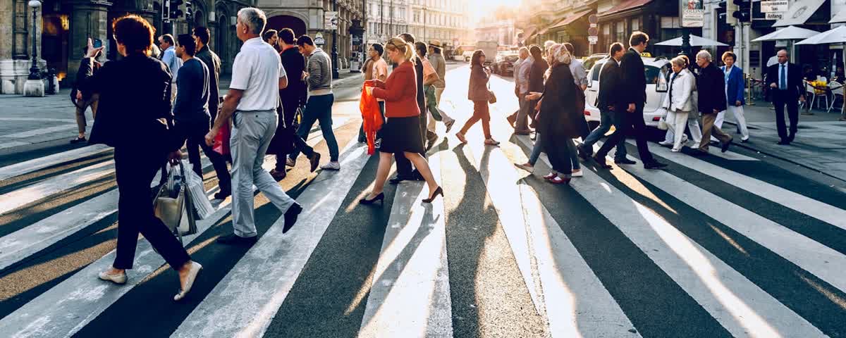 Pessoas a atravessar uma passadeira ao fim da tarde, numa rua movimentada